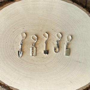 May include: Five silver-toned dangle earrings with lever-back closures. The earrings are shaped like a shovel, razor blade, axe, fish hook, and cleaver. The earrings are displayed on a light brown, wooden surface.