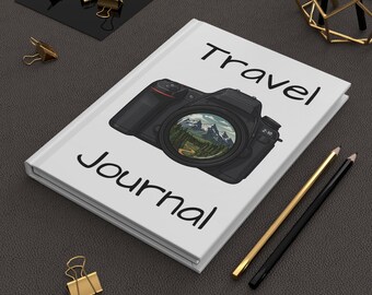 May include: A white Travel Journal with a black camera illustration on the cover. The camera lens shows a mountain landscape. The words "Travel" and "Journal" are printed in black. Gold and black pencils and paper clips are also visible.