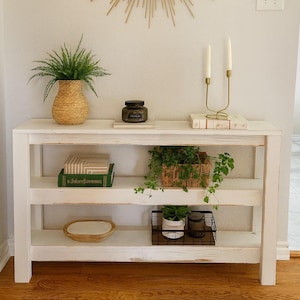May include: A white console table with three shelves, adorned with decorative items. A gold sunburst mirror hangs above. Items include a woven vase with greenery, books, candles, and a wire basket with a plant. The overall aesthetic is modern and minimalist.