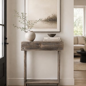 May include: A rustic wooden console table with a framed landscape print, a ceramic vase with greenery, a small bowl, and two books. The table is made of weathered wood and has a lower shelf for storage. The artwork is a neutral-toned landscape.