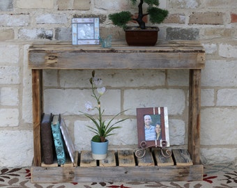 May include: A rustic wooden console table with two shelves. The top shelf has a small framed picture and a potted bonsai tree. The bottom shelf has a stack of books and a framed photo of a couple. The table is made of reclaimed wood and has a natural, weathered finish.