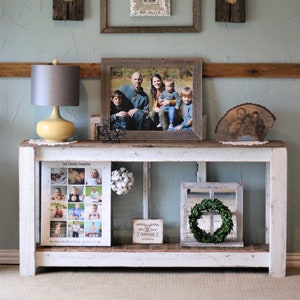May include: A white farmhouse style console table with two shelves. The top shelf has a framed photo of a family, a small wooden sign that says "After all, I'm just a girl, standing in front of a boy, asking him to love her", and a white cotton ball garland. The bottom shelf has a framed photo collage, a framed wreath, and a small wooden sign that says "Farmhouse Vibes".