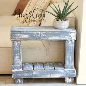 May include: A distressed gray wooden end table with a shelf. The table has a whitewashed finish and is sitting next to a beige couch. A small potted succulent plant is on top of the table.