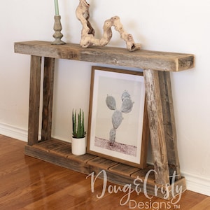 May include: A rustic wooden console table with a distressed finish. The table has a single shelf and two angled legs. The table is decorated with a green candle, a piece of driftwood, a potted succulent, and a framed print of a cactus.