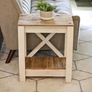May include: White painted end table with a wooden top and a lower shelf. The table has an X-shaped design on the front and is sitting on a white rug with a geometric pattern.
