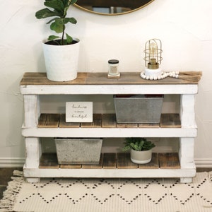 May include: A white wooden shelf unit with two shelves, featuring a potted plant, a small candle, a decorative light fixture, and a sign that reads "Kindness is always beautiful".