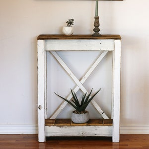 May include: White wooden console table with an X-shaped design on the front. The table has a top shelf and a lower shelf. There are two small potted plants on the top shelf.