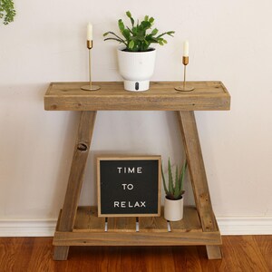 May include: A rustic wooden console table with two shelves. The top shelf has two gold candle holders and a white planter with a green succulent. The bottom shelf has a black letter board with the text "TIME TO RELAX" and a white planter with a green succulent.