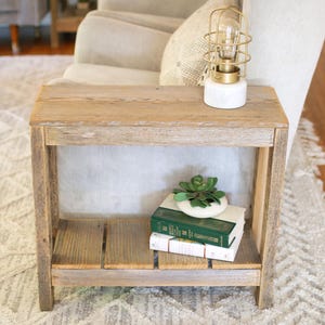 May include: A rustic wooden end table with a single shelf. The table has a natural wood finish and is sitting next to a white couch. A small white lamp with a gold cage is on the table, and a potted succulent plant is on the shelf.