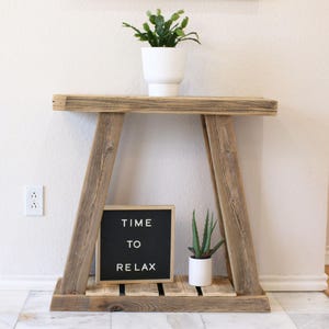 May include: A rustic wooden console table with a distressed finish. The table has two shelves and is decorated with a potted succulent and a letter board that reads "TIME TO RELAX".