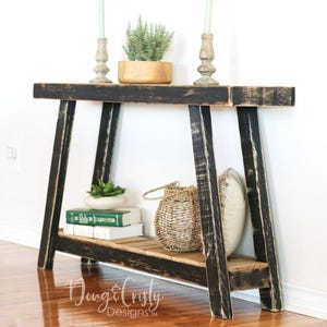 May include: A black wooden console table with a distressed finish. The table has a shelf and two open sides. It is decorated with a potted plant, a woven basket, and a pillow.