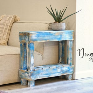 May include: A small, blue and white distressed wood end table with a single shelf. The table is in front of a light beige couch with a throw pillow.