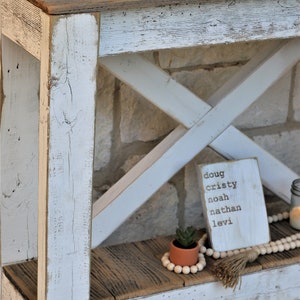 May include: Whitewashed wooden console table with a distressed finish. The table has a shelf and a top made of reclaimed wood. A small potted succulent and a wooden sign with the names Doug, Cristy, Noah, Nathan, and Levi are on the shelf.
