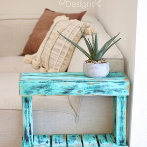 May include: A teal blue end table with a distressed finish and a single shelf. The table is made of wood and has a rustic look. A small potted plant sits on the top shelf.