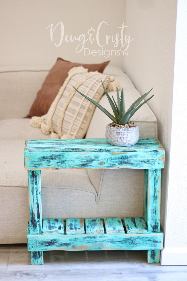 May include: A teal blue end table with a distressed finish and a single shelf. The table is made of wood and has a rustic look. A small potted plant sits on the top shelf.