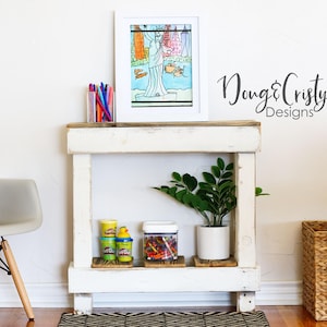 May include: A white wooden console table with a framed artwork, a potted plant, and storage containers. The artwork depicts a colorful illustration. A gray chair with a stuffed animal sits to the left. The text "Doug & Cristy Designs" is visible.