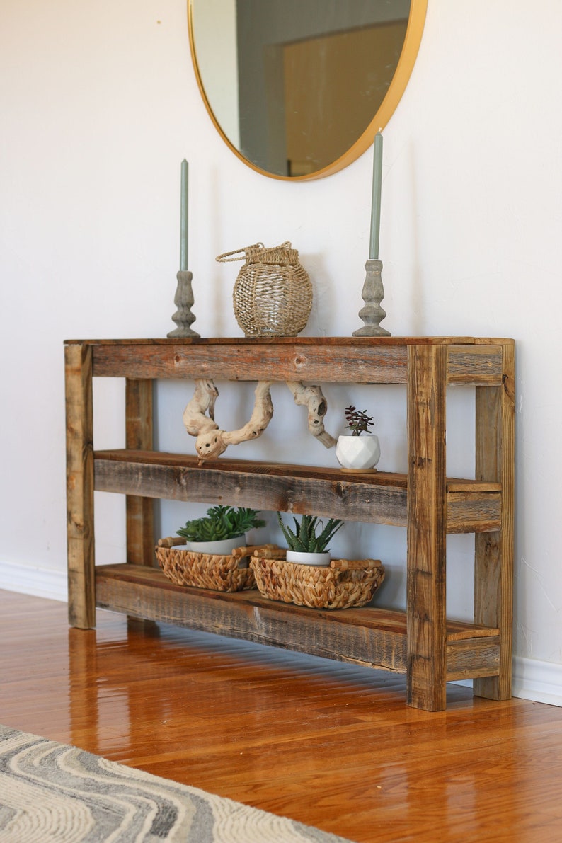 Puede incluir: Una mesa consola de madera r&uacute;stica con dos estantes, con un acabado de madera natural. La mesa est&aacute; decorada con velas, una linterna de mimbre y plantas en macetas.