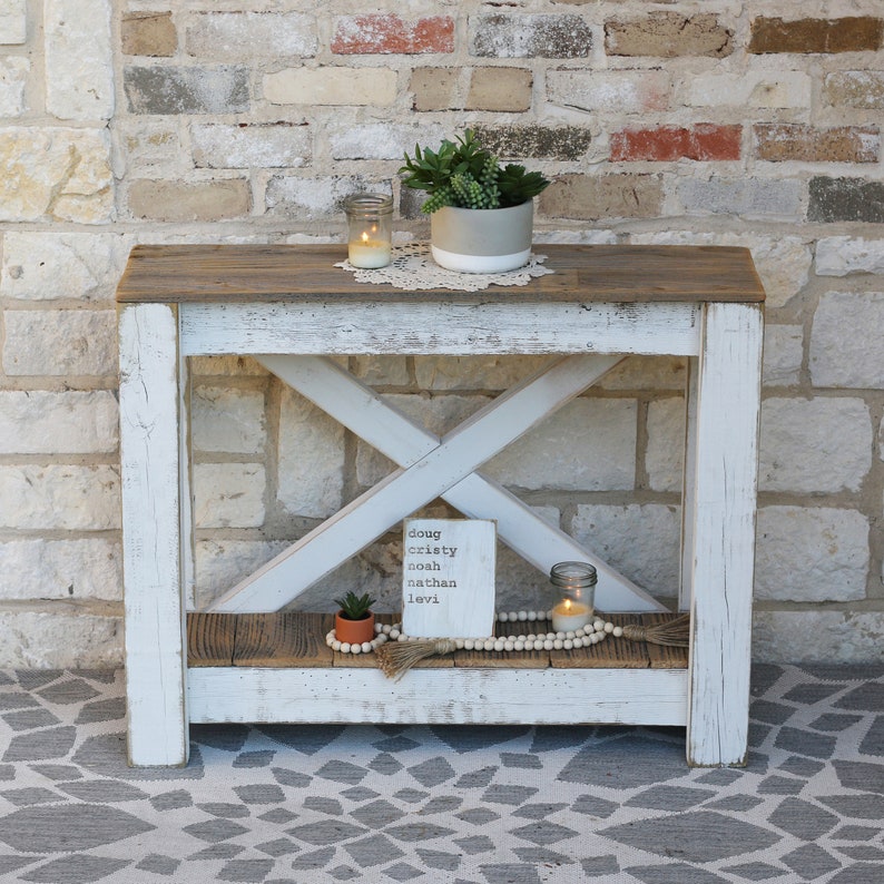 May include: White farmhouse style console table with a distressed wood top and a lower shelf. The table has a whitewashed finish and a criss-cross design on the front. A small potted plant, a candle, and a wooden sign with the names "Doug, Christy, Noah, Nathan, Levi" are on the table.