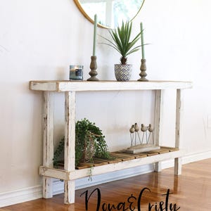 May include: White distressed wood console table with a lower shelf. The table has a rustic farmhouse style and features a distressed white finish. The table is decorated with plants, candles, and decorative birds.