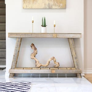 May include: A rustic wooden console table with a natural driftwood accent. The table features a light gray finish, a lower shelf, and is decorated with a small potted plant and two gold candle holders. The table is placed against a white wall.