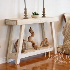 May include: White wooden console table with a distressed finish. The table has two shelves and is decorated with driftwood, a potted plant, and two candlesticks. The table is sitting on a wooden floor.
