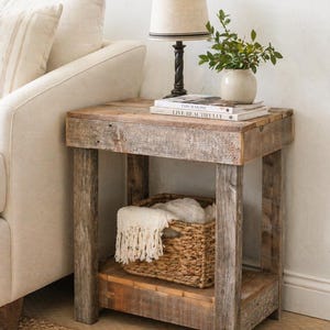 May include: A rustic wooden side table with a lower shelf and a woven basket. A lamp with a beige shade and a dark base sits on top, along with books and a small vase with greenery. The table is next to a beige sofa.