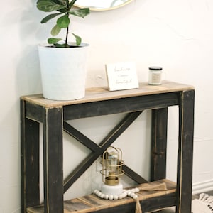 May include: A black wooden console table with an X-shaped design on the bottom shelf. The table has a distressed finish and a light wood top. A small white sign with the text "Kindness is always beautiful" is on the top shelf, along with a white candle and a small gold cage light.