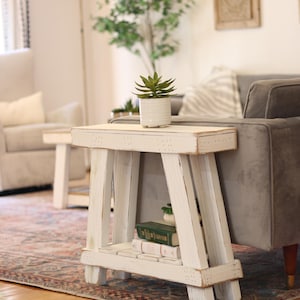 May include: White wooden side table with a distressed finish. The table has a shelf and a top surface. A small potted succulent plant sits on the top surface. The table is in a living room setting with a rug and a couch.