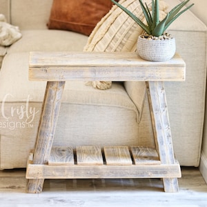 May include: A rustic wooden side table with a distressed white finish. The table has a single shelf and a simple A-frame design. It is sitting in front of a white couch with a plant on top.