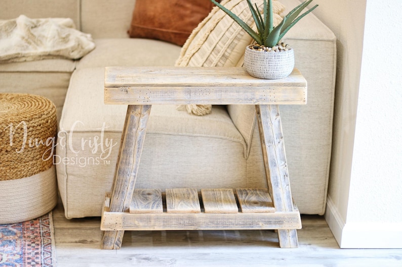 May include: A rustic wooden side table with a distressed white finish. The table has a single shelf and a simple A-frame design. It is sitting in front of a white couch with a plant on top.