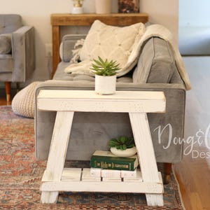 May include: White wooden side table with a distressed finish, featuring a shelf for storage. The table is positioned next to a gray sofa with a white throw blanket and a potted green plant.