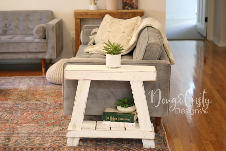 May include: White wooden side table with a distressed finish, featuring a shelf for storage. The table is positioned next to a gray sofa with a white throw blanket and a potted green plant.