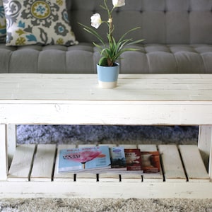 May include: A white, rectangular coffee table with a distressed finish. The table has a lower shelf holding magazines and a small blue and white flower pot on top. The table is in front of a gray couch with decorative pillows.