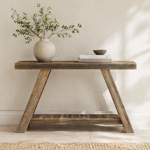 May include: A rustic wooden console table with a natural finish. The table features a lower shelf and angled legs. A round, textured vase with greenery, books, and a small bowl are displayed on the tabletop. The table is approximately 48 inches long.