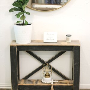 May include: A black wooden console table with an X-shaped design on the lower shelf. The table has a wooden top and a white ceramic plant pot with a green plant. A small white sign with the text "Kindness is always beautiful" is on the table. A small candle and a gold cage light with a white beaded garland are on the lower shelf.
