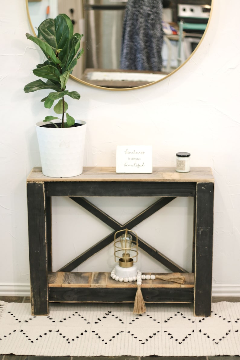 May include: A black wooden console table with an X-shaped design on the lower shelf. The table has a wooden top and a white ceramic plant pot with a green plant. A small white sign with the text "Kindness is always beautiful" is on the table. A small candle and a gold cage light with a white beaded garland are on the lower shelf.