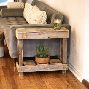May include: A rustic wooden side table with a lower shelf, featuring a distressed white paint finish. The table is next to a gray sofa and a woven basket with a green plant.
