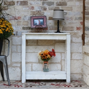 May include: White wooden console table with a shelf, featuring a framed photo, a lamp with a beige shade, and a vase of fall flowers.