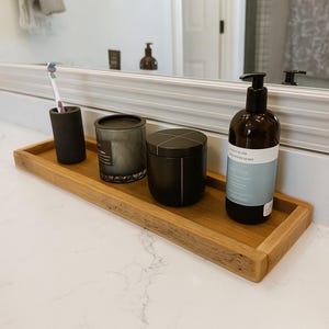 May include: A wooden tray holds a black toothbrush holder, a gray candle, a black container, and a brown bottle with a pump. The bottle has a label that reads "DETOX". The tray sits on a white countertop.