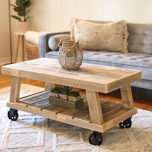 May include: A rustic wooden coffee table with a lower shelf and wheels. The table is made of reclaimed wood and has a natural finish. A woven basket with a handle sits on the top shelf, and a small planter with green plants sits on the lower shelf.