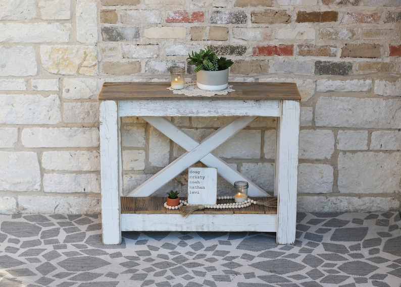 May include: White farmhouse style console table with a distressed wood top and a lower shelf. The table has an X-shaped support structure and is decorated with plants, candles, and a sign that reads "doug okety noah nathan love".