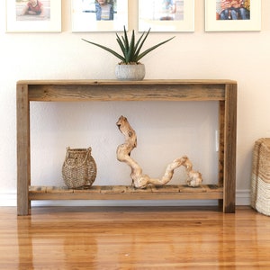 May include: A rustic wooden console table with a lower shelf. The table is decorated with a woven basket, a piece of driftwood, and a potted succulent plant. The table is in front of a white wall with framed pictures.