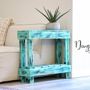May include: A teal blue wooden end table with a distressed finish. The table has two shelves and is sitting on a light brown floor. The table is next to a white wall with the text "Doug & Cristy Designs&trade;" on it.