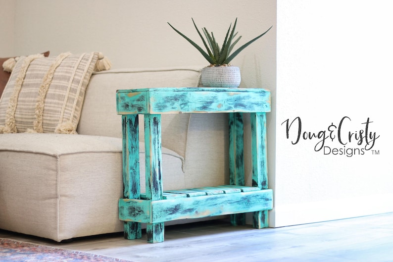 May include: A teal blue wooden end table with a distressed finish. The table has two shelves and is sitting on a light brown floor. The table is next to a white wall with the text "Doug & Cristy Designs&trade;" on it.