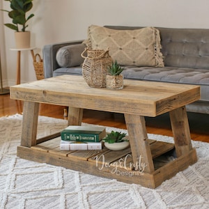 May include: A rustic wooden coffee table with a lower shelf. The table has a natural wood finish and is made of reclaimed wood. The table is decorated with a woven basket, a potted plant, and a stack of books. The table is sitting on a white rug with a geometric pattern.