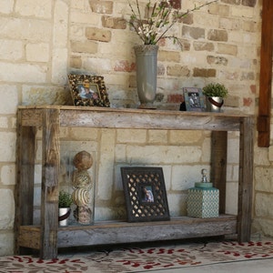 Puede incluir: Una mesa consola de madera r&uacute;stica con dos estantes. La mesa est&aacute; hecha de madera reciclada y tiene un acabado natural. Est&aacute; decorada con un jarr&oacute;n de flores, una foto enmarcada y un tarro decorativo.