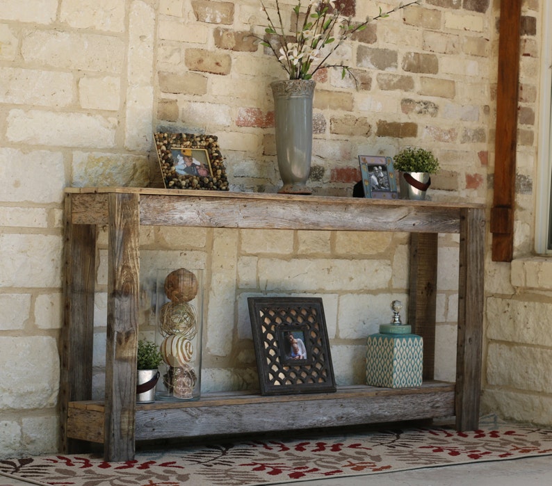 Puede incluir: Una mesa consola de madera r&uacute;stica con dos estantes. La mesa est&aacute; hecha de madera reciclada y tiene un acabado natural. Est&aacute; decorada con un jarr&oacute;n de flores, una foto enmarcada y un tarro decorativo.