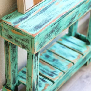 May include: A distressed teal green wooden console table with a shelf. The table has a rustic, farmhouse style and is perfect for displaying decor or storing items.