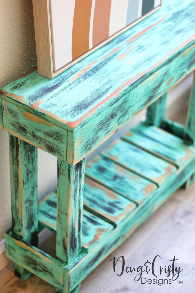 May include: A distressed teal green wooden console table with a shelf. The table has a rustic, farmhouse style and is perfect for displaying decor or storing items.