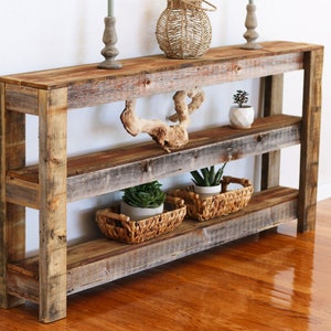 Puede incluir: Mesa consola de madera r&uacute;stica con tres estantes. La mesa est&aacute; hecha de madera recuperada y tiene un acabado natural. Cuenta con una pieza decorativa de madera a la deriva en el estante superior y dos plantas en maceta en el estante inferior.